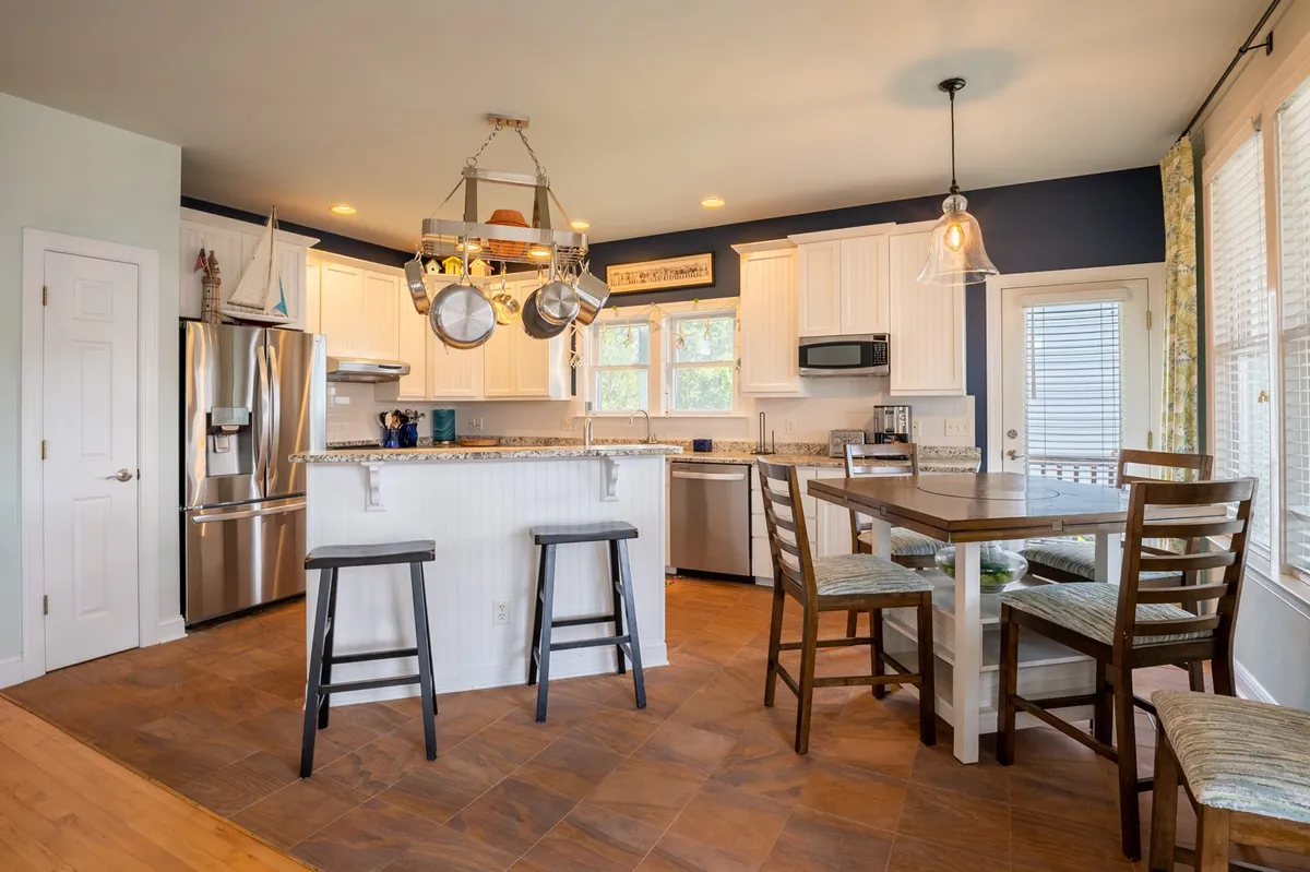 A contemporary kitchen and dining area with a prominent granite countertop island, hanging pots, and a nearby dining table. The space is warm and inviting, clearly designed for cooking, dining, and casual conversations. The kitchen’s layout is open, allowing seamless movement between cooking and social areas.