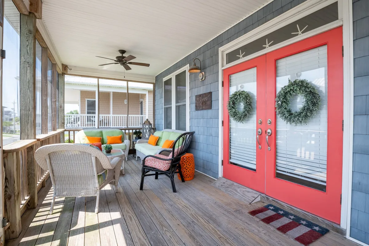 This exterior image features inviting orange double doors adorned with wreaths, colorful cushions on outdoor seating, and a wooden floor, all bathed in bright morning light. The space hints at both comfort and connection to the outdoors, with elements that encourage relaxing and socializing in fresh air.
