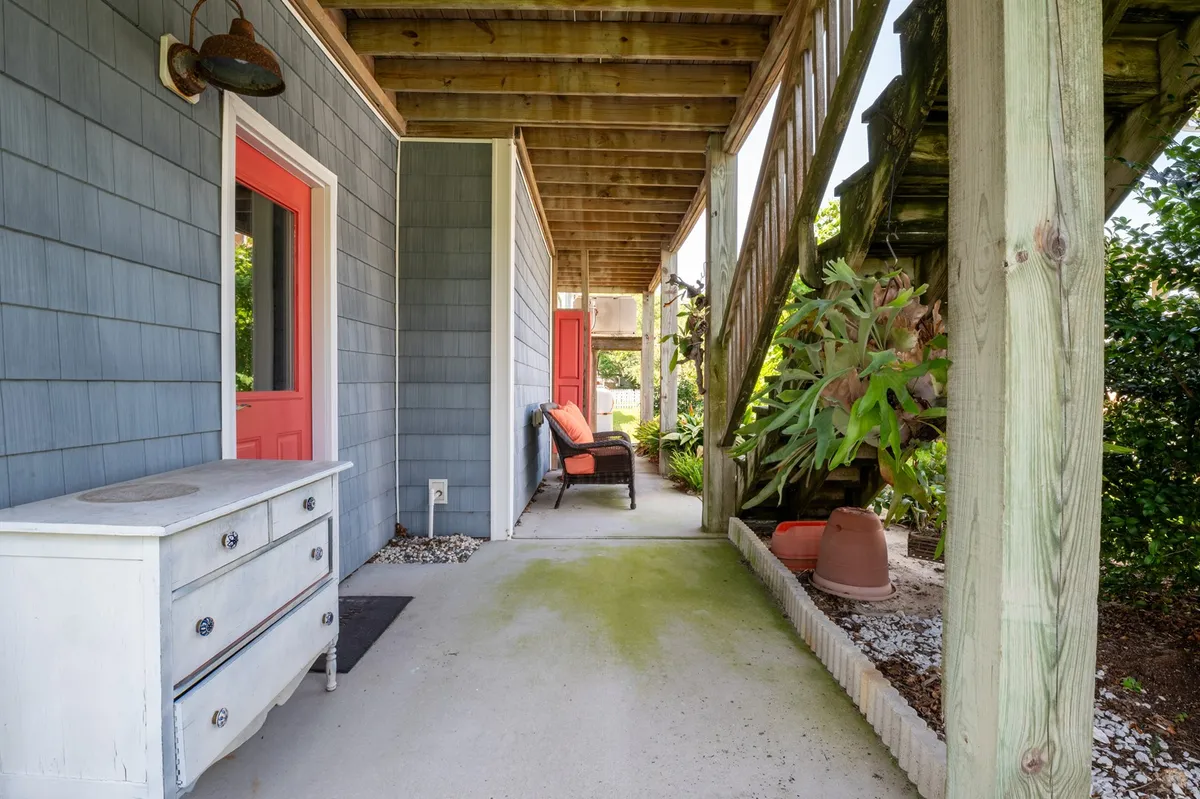 A welcoming porch scene with a red door, white dresser, lush green plants, and a cozy outdoor seating area. Sunlight filters through, inviting guests to relax and enjoy the ambiance just steps from the house.