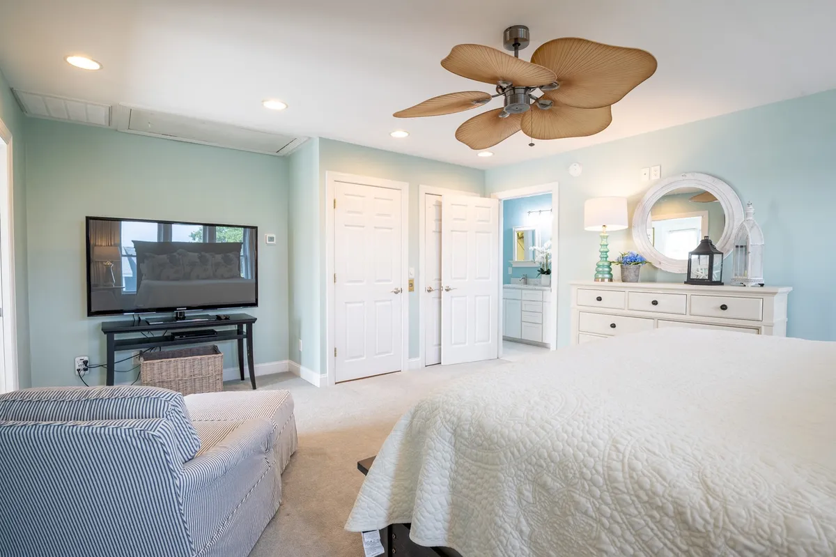 A cozy guest bedroom with a bed, ceiling fan, TV, dresser, and a doorway. The room feels inviting and functional, with natural light adding warmth. The setup emphasizes comfort, convenience, and modern amenities.