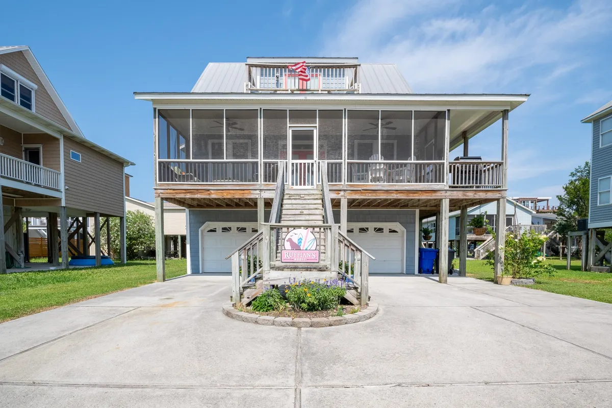 The image showcases the charming exterior of a single-family house at Carolina Beach. The front steps invite guests up to a spacious porch adorned with an American flag, large windows fill the upper floor with light, and a decorative garden sign adds a touch of personalized hospitality. The scene is sunny and welcoming, hinting at the possibilities for porch gatherings, relaxing in the garden, and enjoying panoramic canal views.