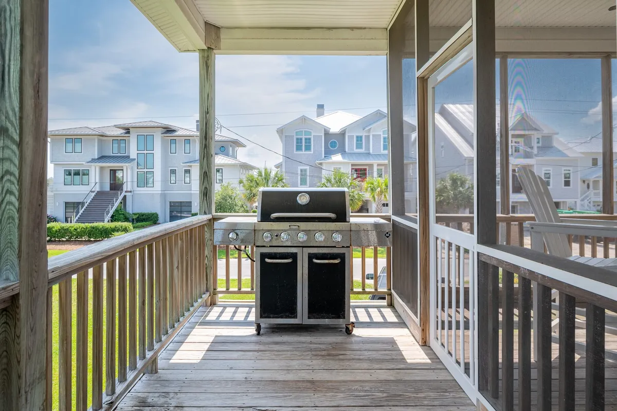 An exterior deck scene featuring a grill, outdoor dining area, and a view of surrounding houses. The setup is ideal for barbecuing, socializing, and enjoying meals while watching the sun set over the waterway.