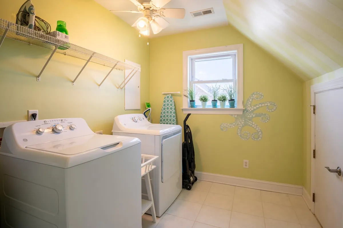 A well-lit laundry area featuring washing machines, potted plants on the window sill, a shelving unit, and cheerful decor. The space is practical yet inviting, with plenty of room for routine chores.