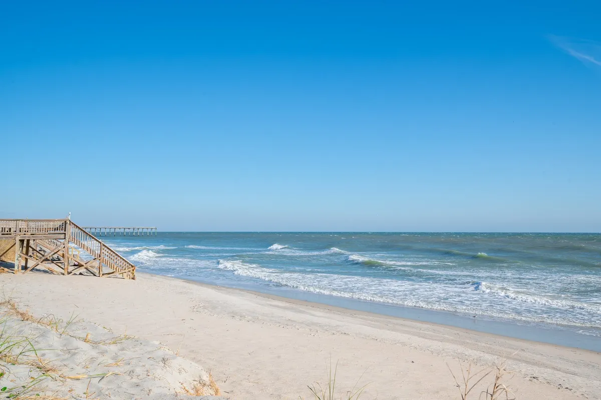 This image captures a sun-soaked beach scene with gentle waves, wooden stairs leading to the sandy shore, and a pier in the distance. The view evokes the excitement and accessibility of Carolina Beach just steps away from the property.