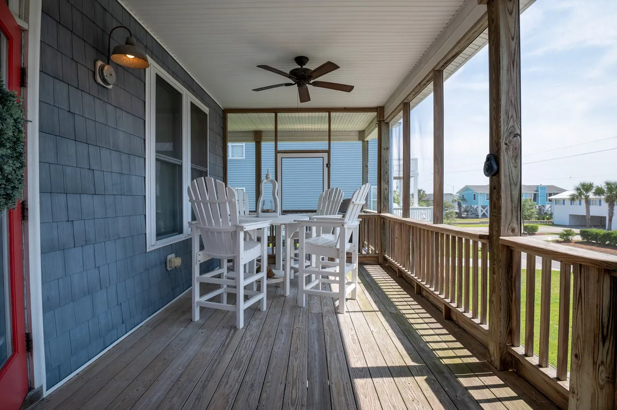A spacious screened porch with a white wooden table and chairs overlooks a tranquil garden and features a blue house exterior. The porch is elevated and surrounded by a wooden railing, offering a comfortable spot for outdoor dining, relaxing with a book, or enjoying coffee while taking in the natural surroundings.