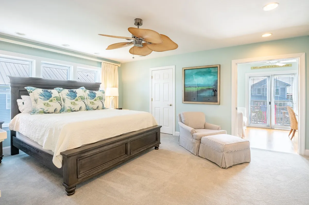 A spacious, serene bedroom featuring a large bed with decorative pillows, a ceiling fan, and an armchair. Natural light flows in, and a doorway leads to another room, suggesting easy access between spaces. The setting is tranquil and welcoming, perfect for restful retreats.
